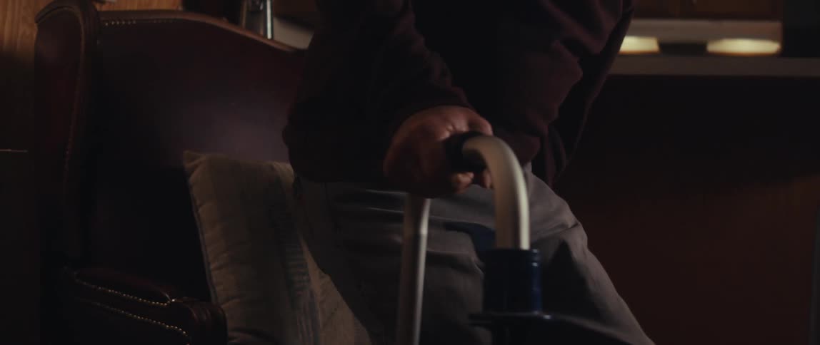 An elderly man with a walker slowly sits on a chair in a dimly lit living room