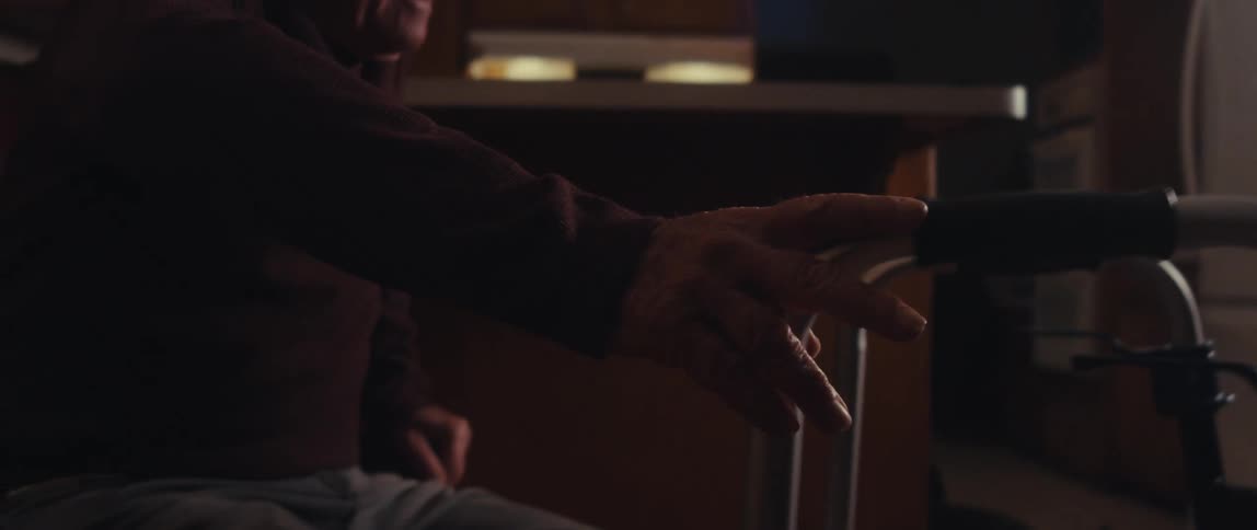 A man with a maroon shirt and gray pants is seen using a walker then reaching and then sitting