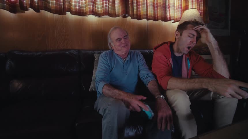 Father and son are sitting on a dark leather couch in a dimly lit living room playing video games