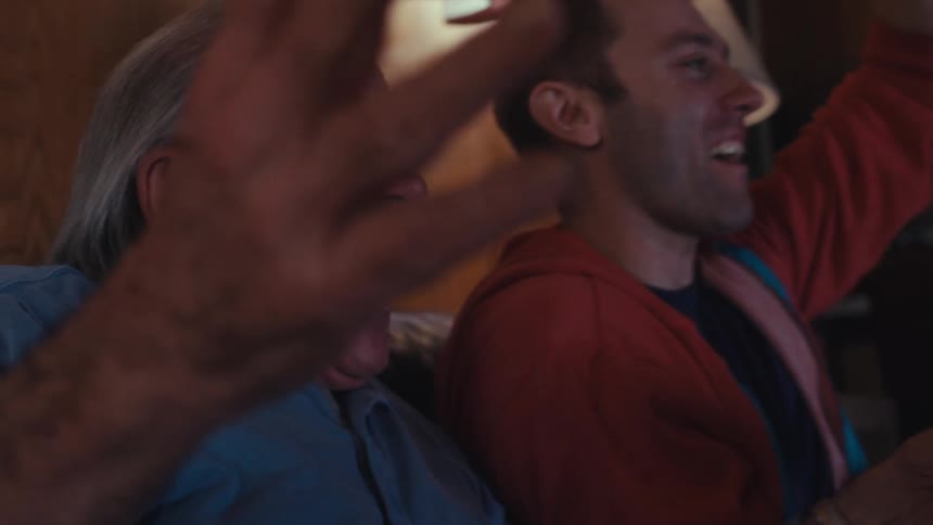 A young man next to an elderly man who are playing a video game and sitting on the couch and then they celebrate and do a high five