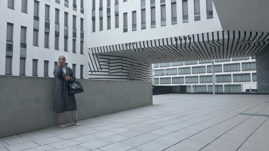 A woman wearing a gray overcoat talking on her cell phone next to a small wall on the outside of a building