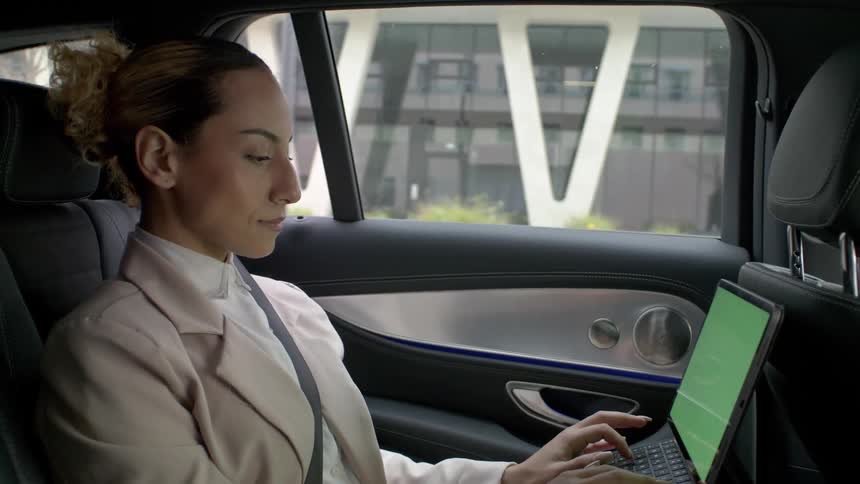 A woman sitting in the back seat of a taxi is working on a laptop as the vehicle drives through the city