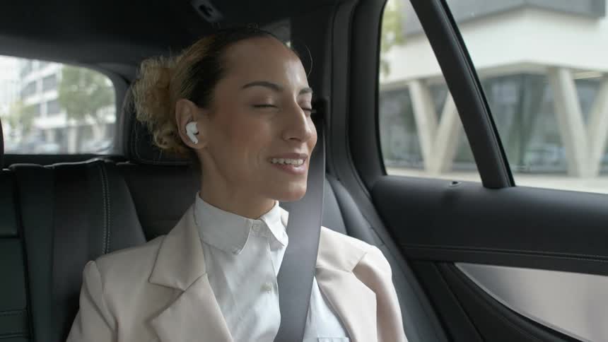 A woman in a light blazer and white shirt wearing earbuds talks to someone while riding in the back of a car