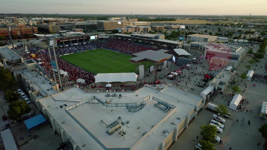 An outdoor stadium filled with spectators during a soccer game surrounded by bustling crowds buildings and parking lots
