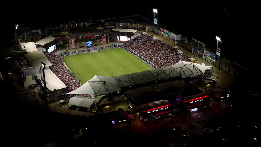 Vehicles drive near a stadium filled with sports fans attending a soccer game at night