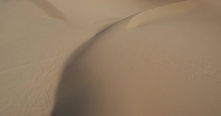 A low aerial shot travels over rolling sand dunes with visible footprints