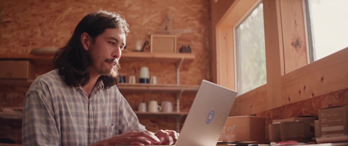 A person is working on a laptop in a wooden room with shelves next to a window allowing natural light to enter