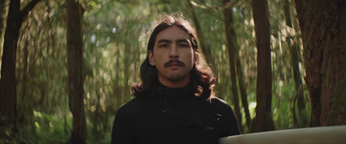 Portrait of a man with long hair and a mustache standing in a forest wearing a black wetsuit