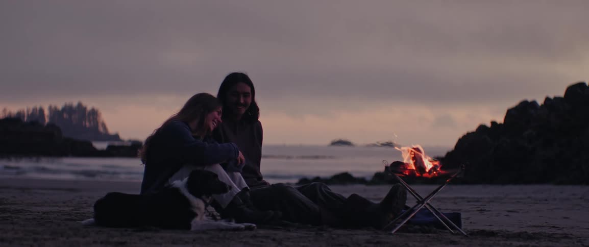 A couple sitting on a beach laughing with each other around a small fire