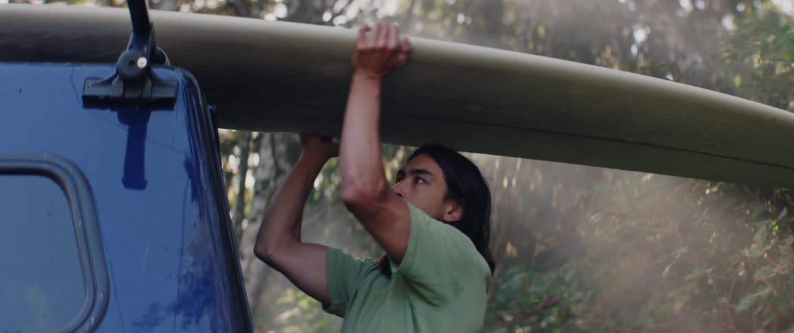 A person unloads a surfboard from the roof of their truck in a forested area
