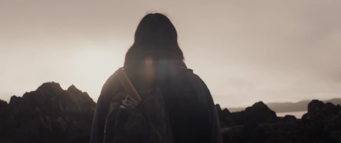 Person with backpack walks on the beach towards a rocky outcrop at sunset