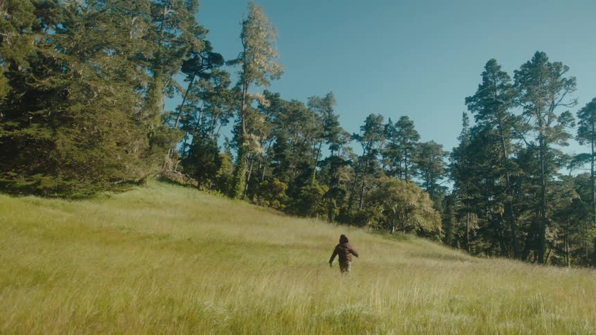 The man in the brown jacket is running through a field of tall grass towards the tree line