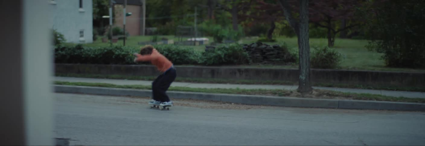 A person in an orange hoodie and dark pants rides a skateboard on a quiet street performing a few basic tricks