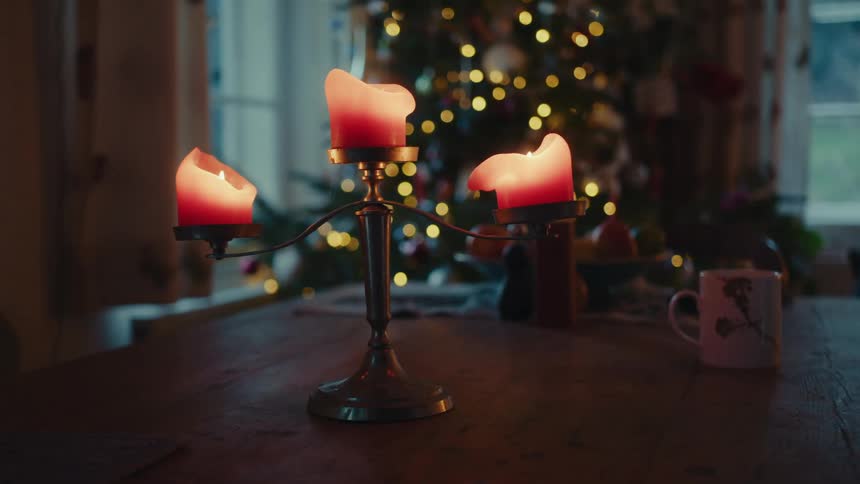 Warm candlelight illuminates a cozy scene casting a soft glow on a wooden table where a candelabra holds three flickering candles while in the background a beautifully decorated christmas tree twinkles with festive lights creating a magical atmosphere