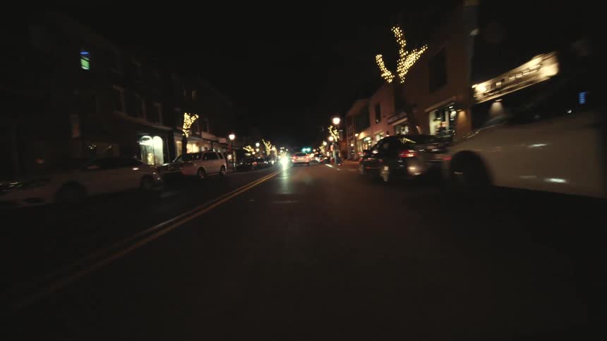 Vehicles and pedestriansmove past stores on a street decorated for christmas at night