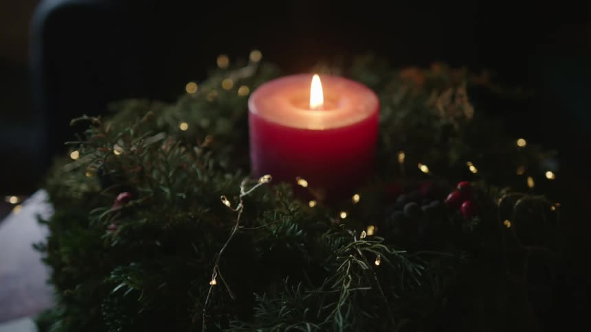 A lit red candle with an orange flame and underneath there are some pieces of plant decorations with twinkling lights