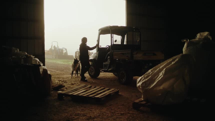 A farmer accompanied by a couple of dogs boards a small truck parked inside a shed on the farm