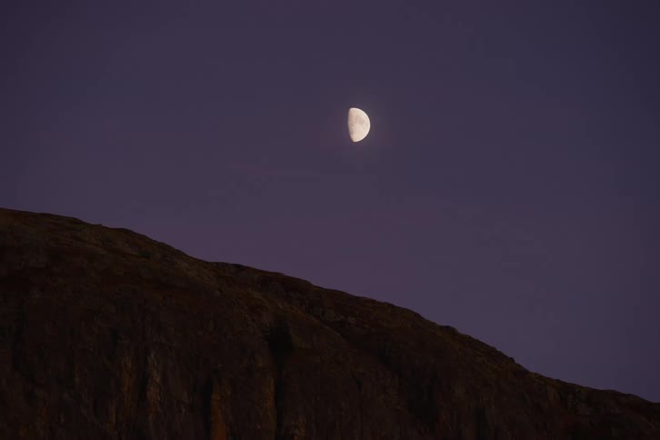 A crescent moon illuminates the darkening sky casting a soft glow over the landscape while a mountain silhouetted against the twilight sky creates a dramatic scene
