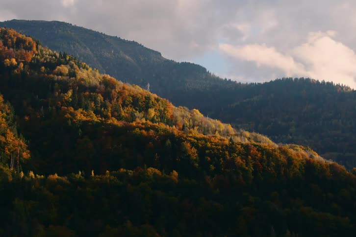 A scenic view of a mountainous landscape with dense forests with a mix of green yellow and orange autumn foliage under a partly cloudy sky