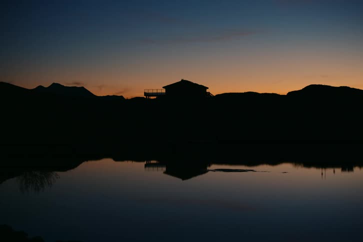 A tranquil scene at dusk where a house stands in silhouette against a vibrant sunset sky its reflection mirroring the beauty of the moment on the still surface of the lake
