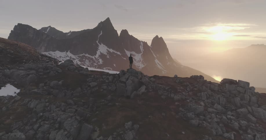 A lone figure standing on a rocky peak silhouetted against a sunset with a mountain range stretching into the distance