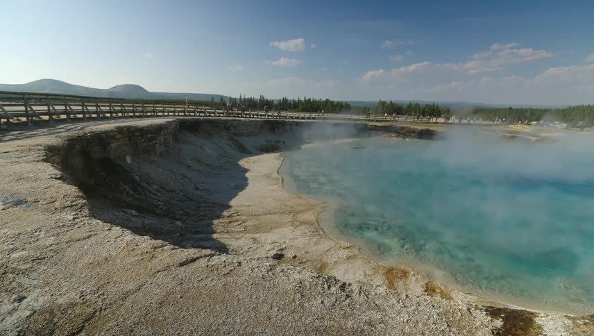 The water in the crater like hole is steaming and there are people walking around the outside of the wooden railing