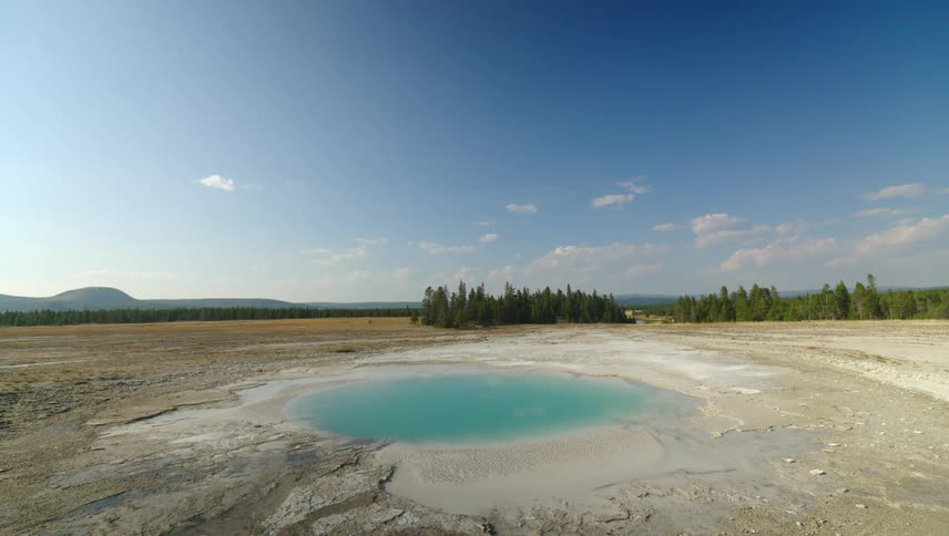 A small vibrant blue hot spring surrounded by a barren landscape under a clear blue sky