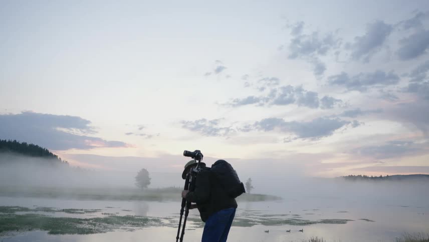 A person wearing a hat is standing with his back turned and holding a camera on a tripod and then takes the camera and leaves in an area with a large lake with fog