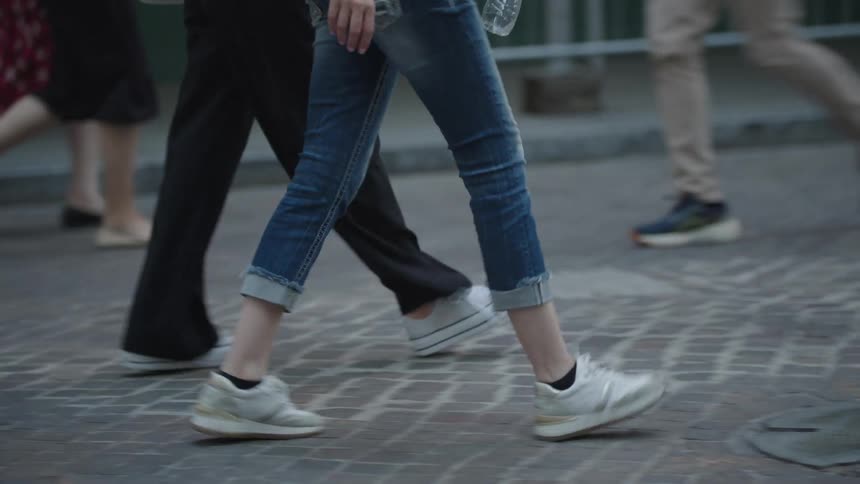 The legs of a person walking on a sidewalk full of pedestrians and that person is wearing white sneakers