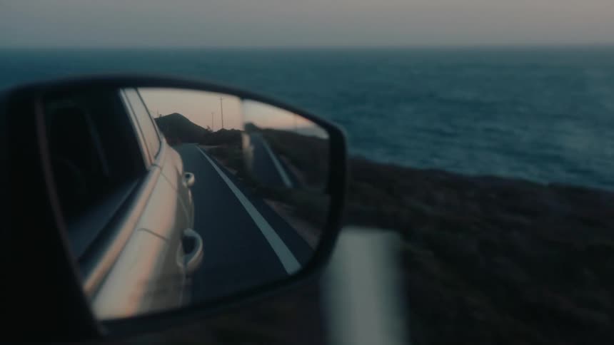 The reflection in a side view mirror shows a winding coastal road disappearing into the distance