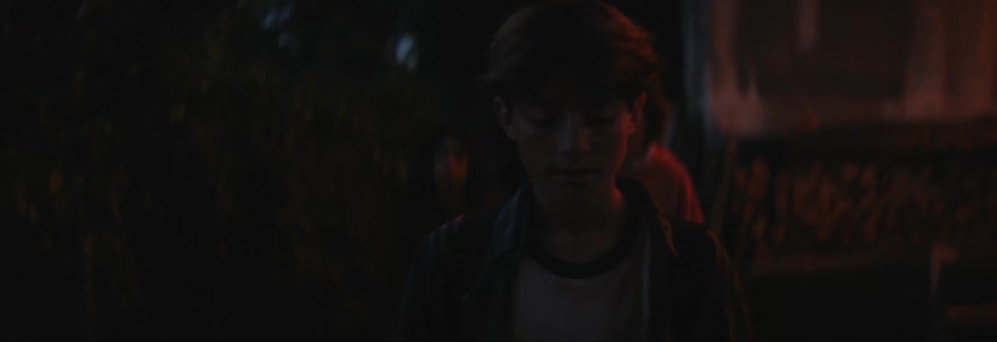A group of teenagers walk under a bridge at night with one of them throwing a skateboard on the ground in frustration