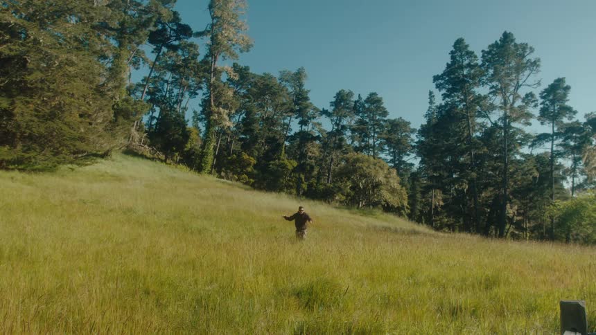 A person is running and jumping through a grassy field surrounded by tall trees under a clear blue sky