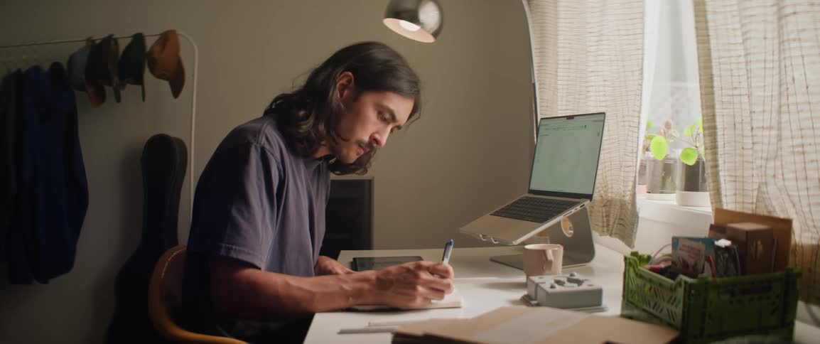 A man writes in a notebook while also working at a tablet and a laptop on a desk