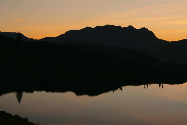Silhouette of people reflecting on a still lake over a mountain range against an orange sunset sky
