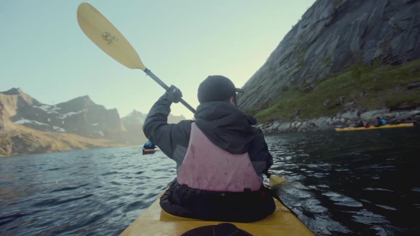 A man wearing a pink life jacket a dark jacket and a cap paddles a yellow kayak across a clear lake surrounded by a scenic landscape of tall rocky mountains with patches of snow and greenery all bathed in the bright sunlight