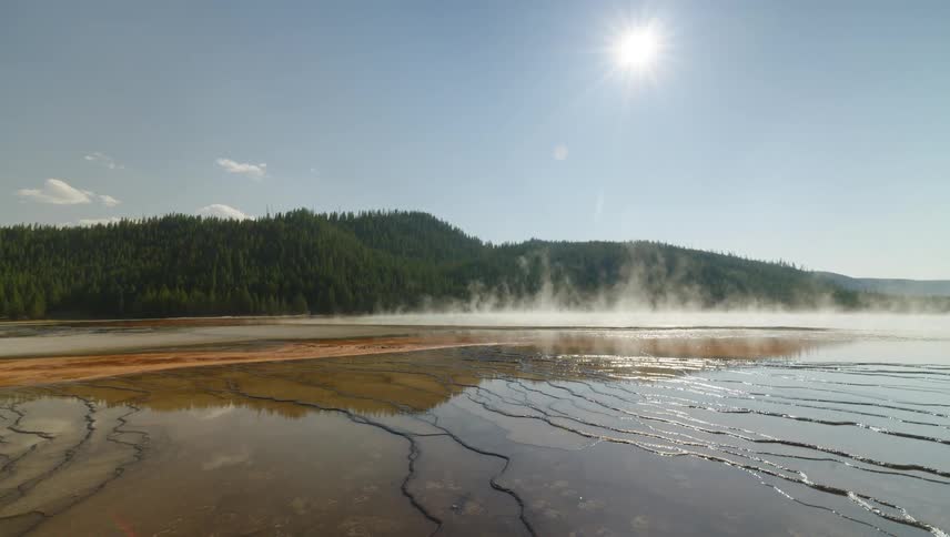 A geothermal spring with colorful mineral deposits steams under a bright blue sky