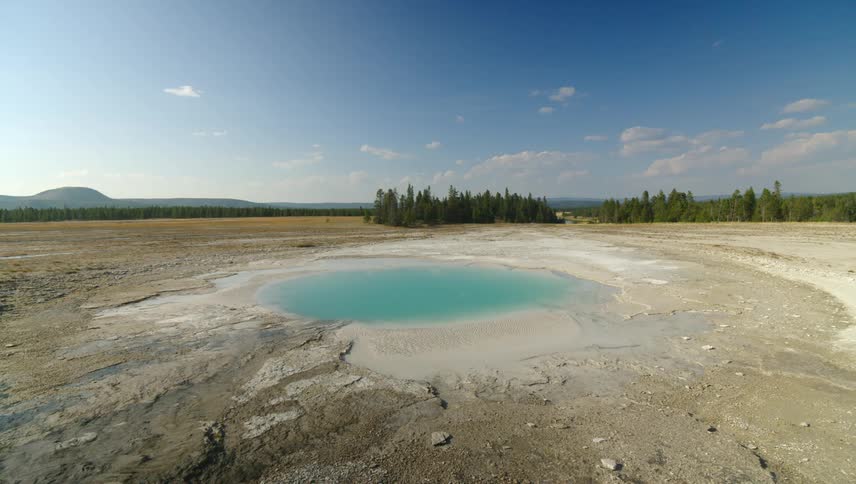 A turquoise hot spring is surrounded by a barren mineralrich landscape with a forested under a clear blue sky