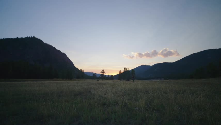 A lone figure walks through a grassy field surrounded by silhouetted mountains as the sun sets