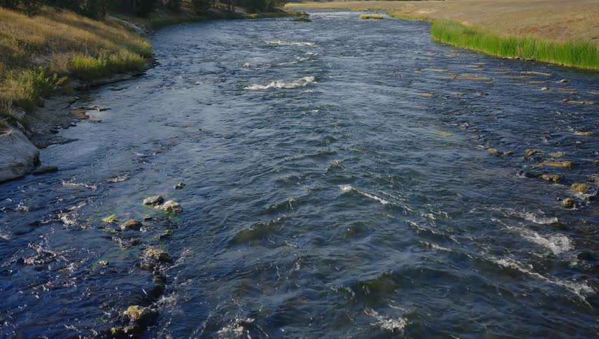 River water flows over the rocks while passing a grassy slope and a field during the daytime