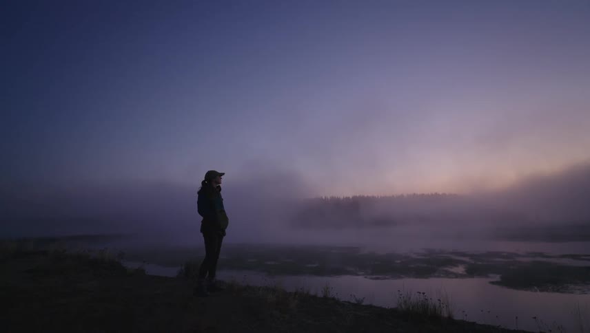 The silhouette of a person wearing a cap standing on a small hill looking ahead where there is a lake and a blue sky