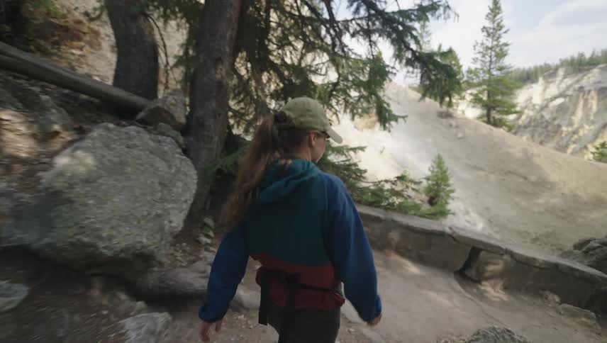 A woman with long hair wearing a cap and a blue jacket is walking on a forest trail surrounded by tall pine trees and large rocks