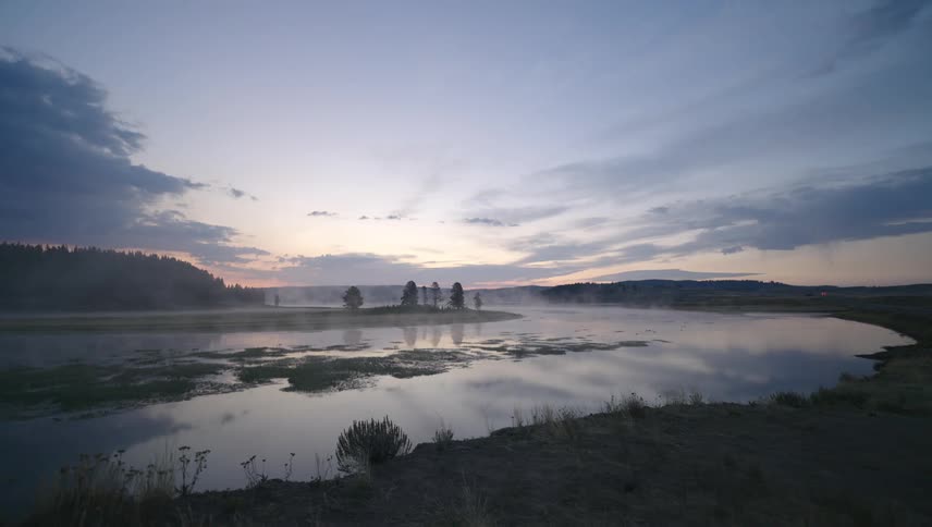 A lakeside scene at dusk with mist rising from the water silhouetted trees on the horizon and a sky with soft hues of blue and pink