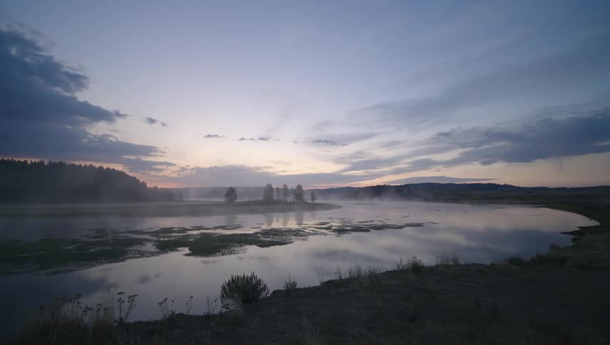 Sun rises on the horizon near a conifer forest bordering a river under a cloudy sky