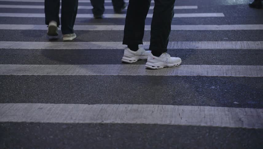 Person in black pants and white sneakers standing in middle of crosswalk as people walk by