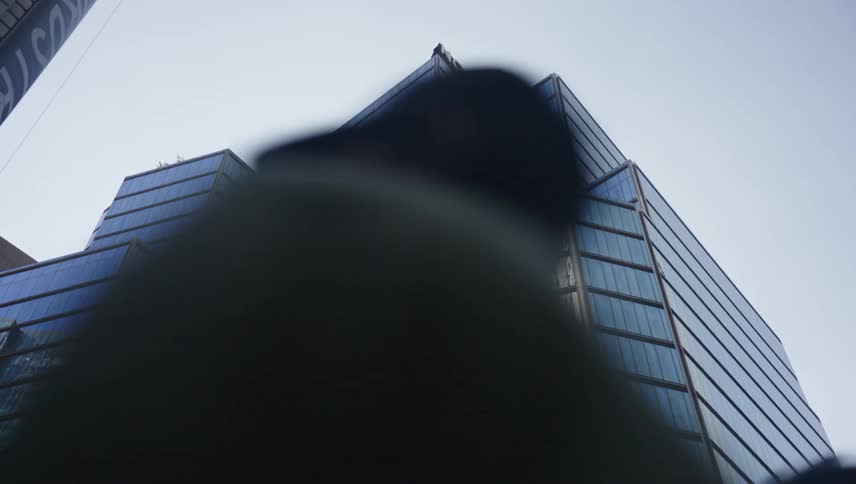 People walk past a modern skyscraper with a reflective glass facade which reflects both the surrounding cityscape and the passersby themselves