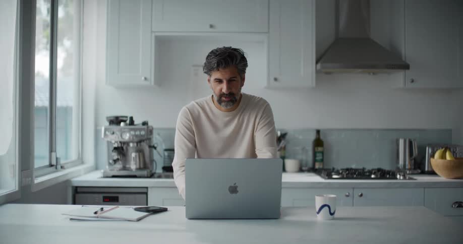 A man with a beard and a cream sweatshirt has a video call on his laptop at the kitchen island with cupboards visible in the background