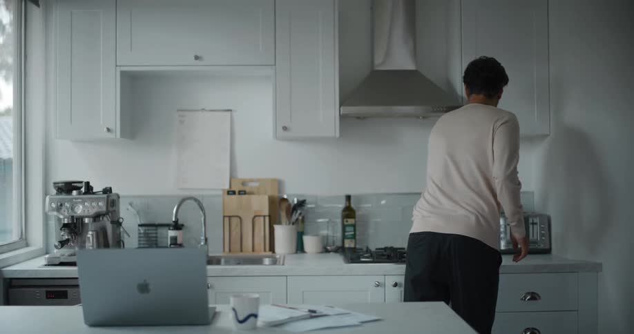 A man works from home with a laptop on the kitchen counter and stands up to make coffee