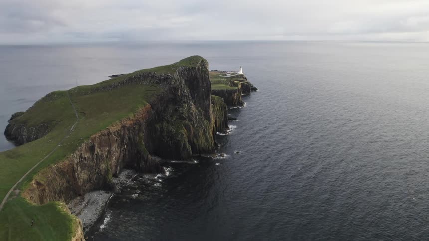 A lighthouse stands dramatically on the edge of a coastal cliff overlooking a vast calm ocean under a cloudy sky