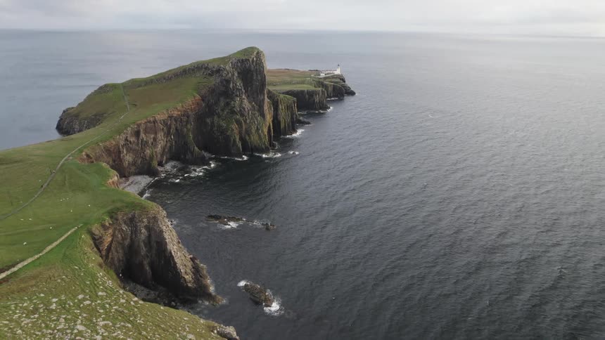 A lighthouse stands at the edge of a dramatic cliff overlooking a vast expanse of the ocean with a winding path leading towards it along the rugged coastline