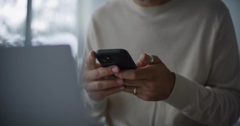 Bearded man in a cream sweatshirt uses a smartphone and a laptop at the kitchen island